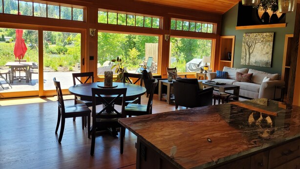 Amplia sala de estar con techo de madera, ventanas grandes que ofrecen vista a la vegetación, mesa de comedor con sillas y un sofá con cojines.