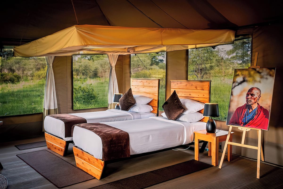 A tented room with two beds, a wooden headboard, a table with a lamp, and a painting on the wall.
