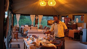 Un área de comedor cubierta con carpas, una mesa lista para dos personas, un hombre sirviendo vino y vistas al exterior a través de las ventanas.