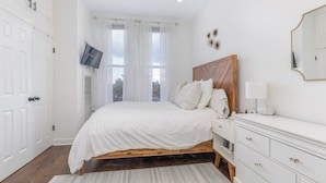 A bedroom with a bed, a TV mounted on the wall, a dresser, and a window with curtains.