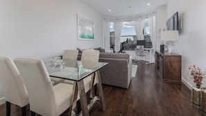 A modern living room with a glass dining table, a sofa, and a TV mounted on the wall.