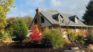 Une maison avec un toit en pente, une façade en briques et un jardin où poussent différentes plantes, ainsi qu’un arbuste à fleurs rouges.