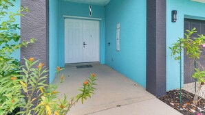 Une entrée d’immeuble avec une façade bleue, une porte blanche et un petit jardin agrémenté de plantes.