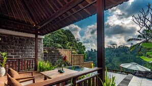 An outdoor patio with wooden furniture, a stone wall, and a view of lush greenery. - Belas Vana Villa Ubud by Dhananjaya Hospitality (Sebatu)