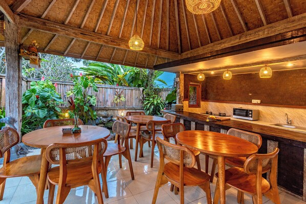 Uma área de refeições de madeira com mesas e cadeiras redondas, um bar com micro-ondas e vista para vegetação exuberante.