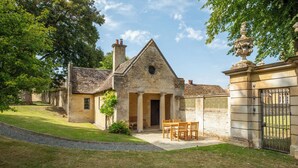 A stone building with a triangular roof, a central entrance with columns, and a small garden with a table and chairs. - The Gate House (Apethorpe)