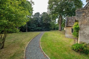 A gravel pathway leading to a stone building with a tiled roof, surrounded by lush greenery and trees. - The Gate House (Apethorpe)