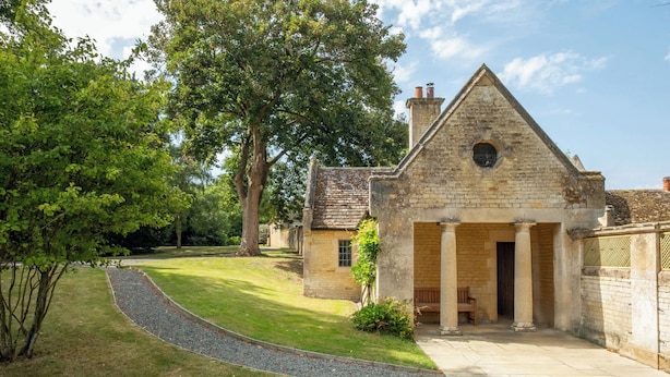 A stone building with a triangular roof and a central doorway, surrounded by a well-maintained lawn and trees. - The Gate House (Apethorpe)