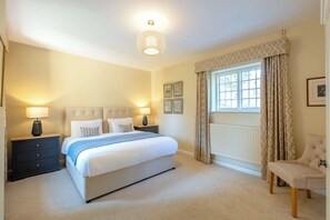 A bedroom with a large bed, two bedside tables with lamps, a chair, a window with curtains, and a radiator. - The Gate House (Apethorpe)