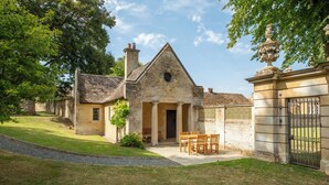 A stone building with a triangular roof, a central entrance with columns, and a small garden with a table and chairs. - The Gate House (Apethorpe)