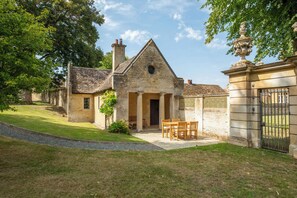 A stone building with a triangular roof, a central entrance with columns, and a small garden with a table and chairs. - The Gate House (Apethorpe)