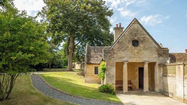 A stone building with a triangular roof and a central doorway, surrounded by a well-maintained lawn and trees. - The Gate House (Apethorpe)