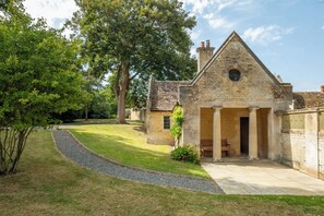A stone building with a triangular roof and a central doorway, surrounded by a well-maintained lawn and trees. - The Gate House (Apethorpe)