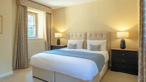 A bedroom with a large bed, two bedside tables with lamps, a window with curtains, and a radiator. - The Gate House (Apethorpe)
