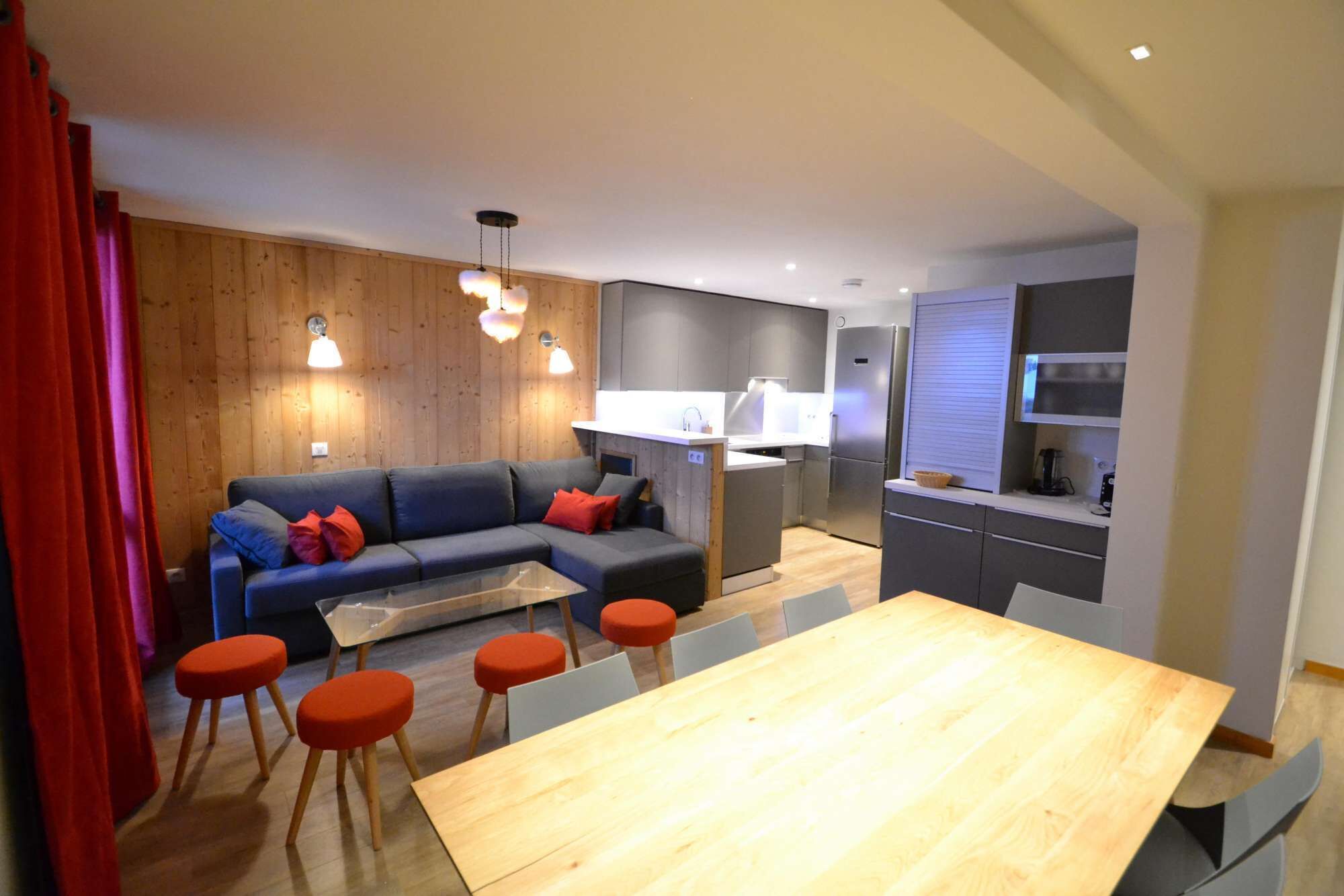 A modern living space with a wooden accent wall, a grey sofa with red cushions, a wooden dining table with red stools, and a kitchen area with grey cabinets and a built-in oven.