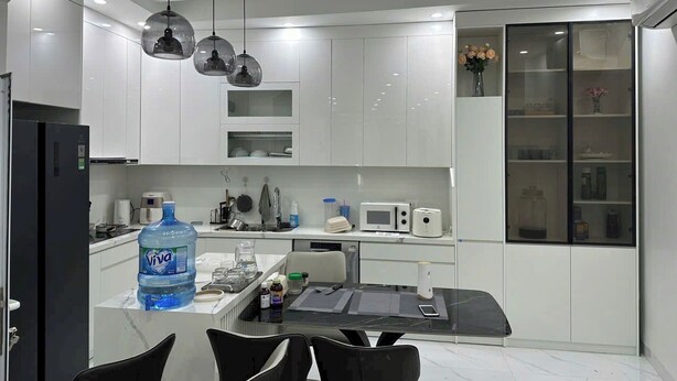 A modern kitchen with white cabinets, a large water dispenser, a microwave, and a glass-fronted cabinet with decorative items. - Oanh Luxstay (Bac Hong Linh)