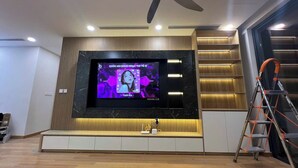A modern living room with a large flat-screen TV mounted on a wooden wall, a ceiling fan, and a staircase with a metal ladder. - Oanh Luxstay (Bac Hong Linh)