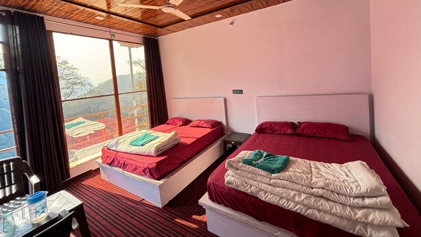 A room with two beds, a ceiling fan, a window with a view of the outdoors, and a table with a water jug. - Kedar Hill Resort  (Ukhimath)
