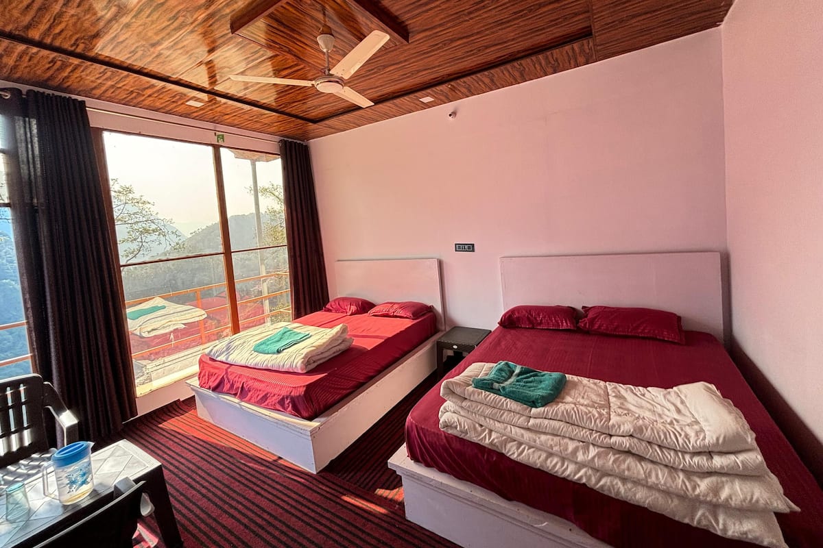 Una habitación con dos camas, un ventilador de techo, una ventana con vistas al exterior y una mesa con un jarro de agua.