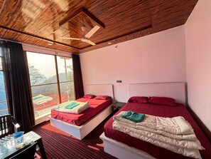 A room with two beds, a ceiling fan, a window with a view of the outdoors, and a table with a water jug. - Kedar Hill Resort (Ukhimath)