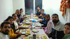 A group of people seated around a long table with various dishes, in a room with a window and a wall-mounted rack of items. - Kedar Hill Resort  (Ukhimath)
