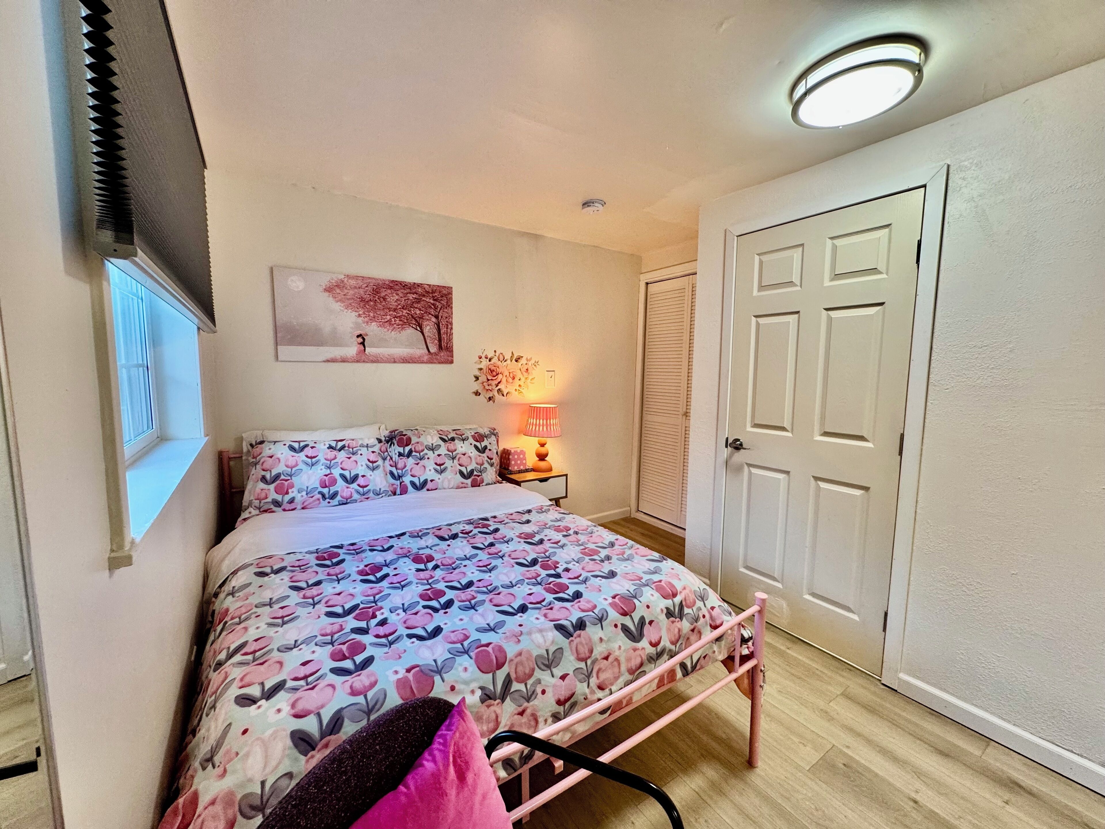 Een slaapkamer met een bed, nachtkastje, lamp, raam, deur en een plafondlamp.