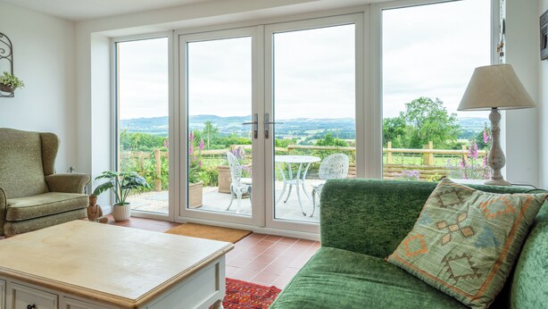 A living room with a green sofa, a white armchair, a small table, and a view of a garden through large sliding glass doors. - Modern Snitton Cottage & Gardens (Snitton)