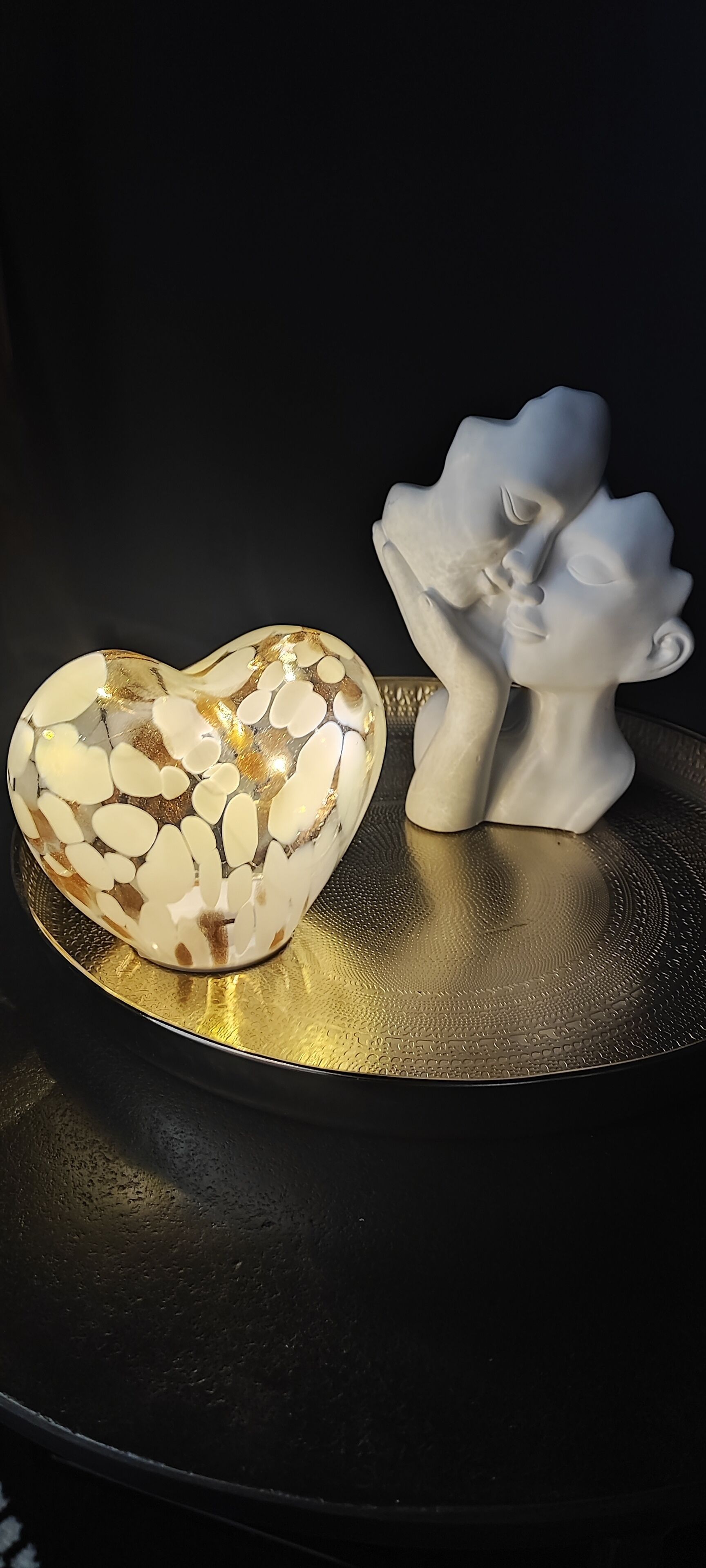 A heart-shaped object with a gold and white pattern on a reflective surface, accompanied by a white sculpture of a face.