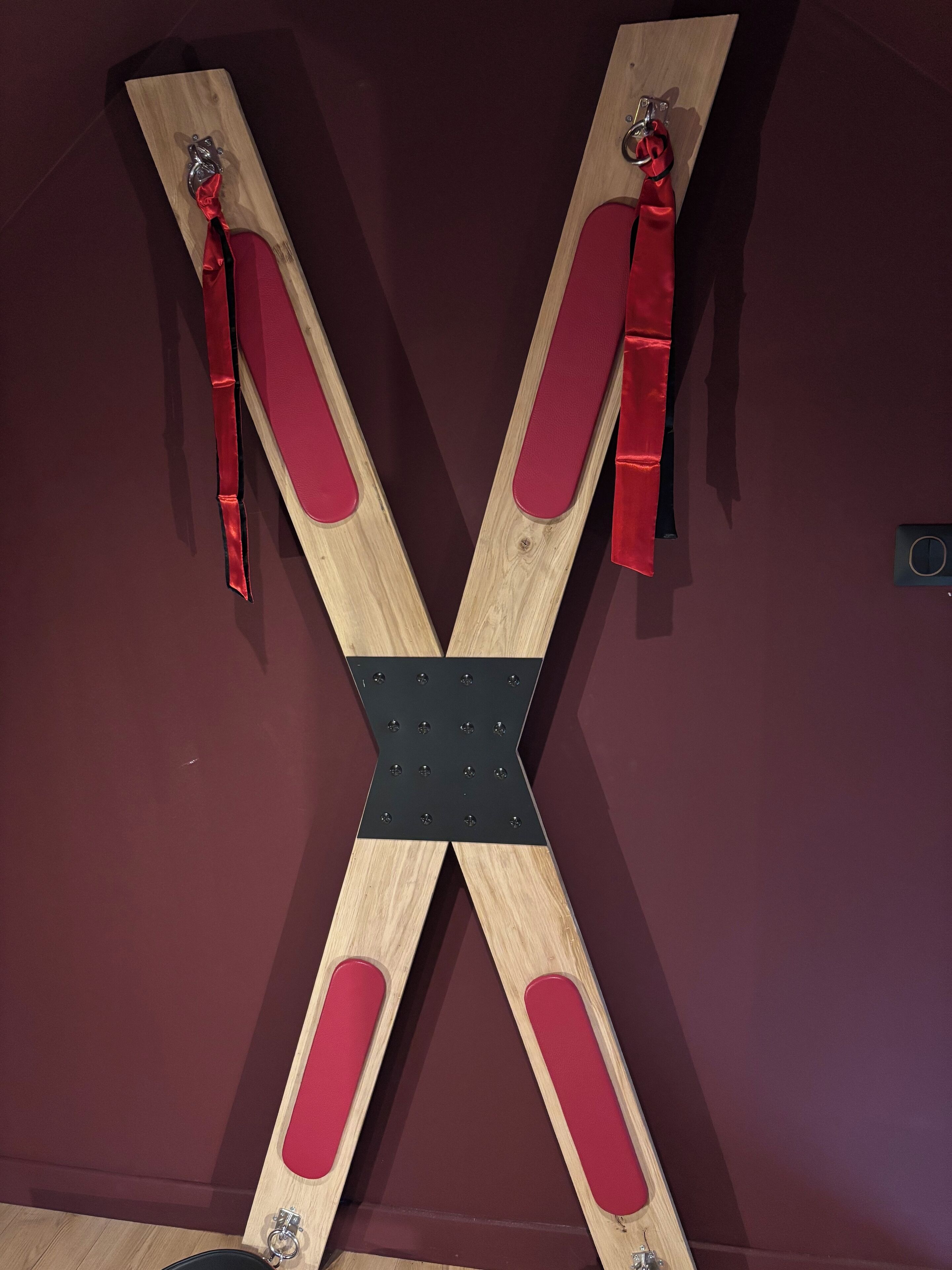 Une structure en bois en forme de X, dotée d’un rembourrage rouge et d’éléments de fixation en métal, fixée à un mur.