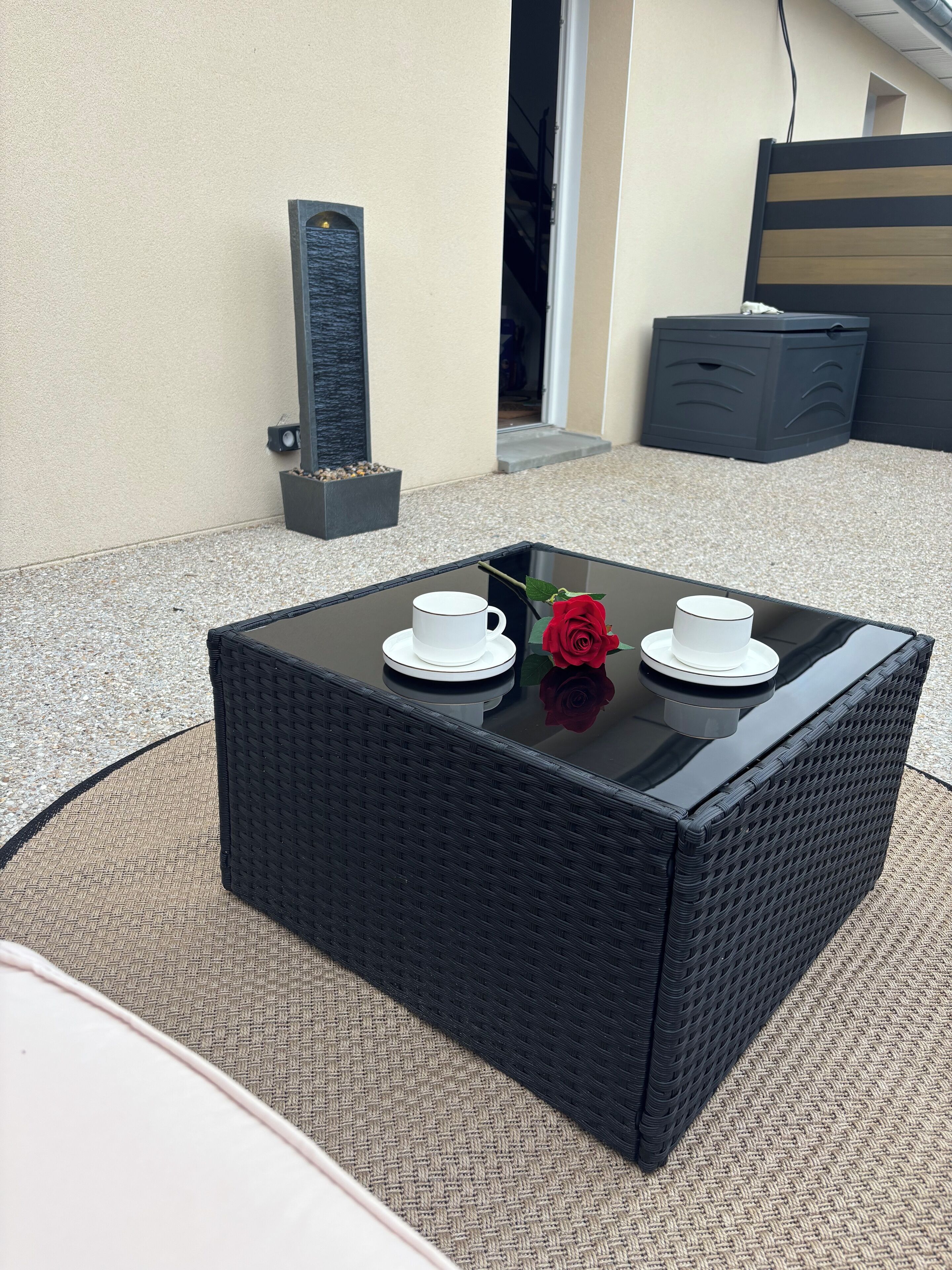 A square outdoor table with two white cups and saucers, a red rose, and a small planter with pebbles.