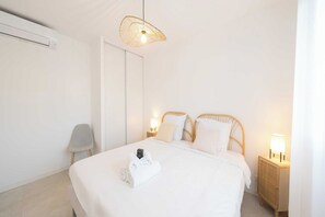 Ein ordentlich bezogenes Bett mit einem gefalteten Handtuch, einer Uhr, einer Nachttischlampe und einem Weidenbeistelltisch.
