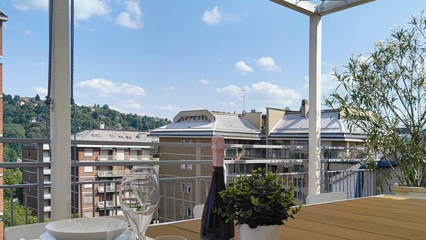 A table set for two with a bottle of wine, glasses, and a bowl on a terrace with a view of buildings and trees. - Piano 7 | Bright penthouse with terrace (Torino)