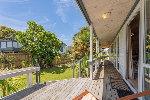 A wooden deck with a railing, a covered porch, and a view of a backyard with trees and a fence. - 5 minutes walk to beautiful beach & shops, fully fenced section pet friendly. (Matarangi)
