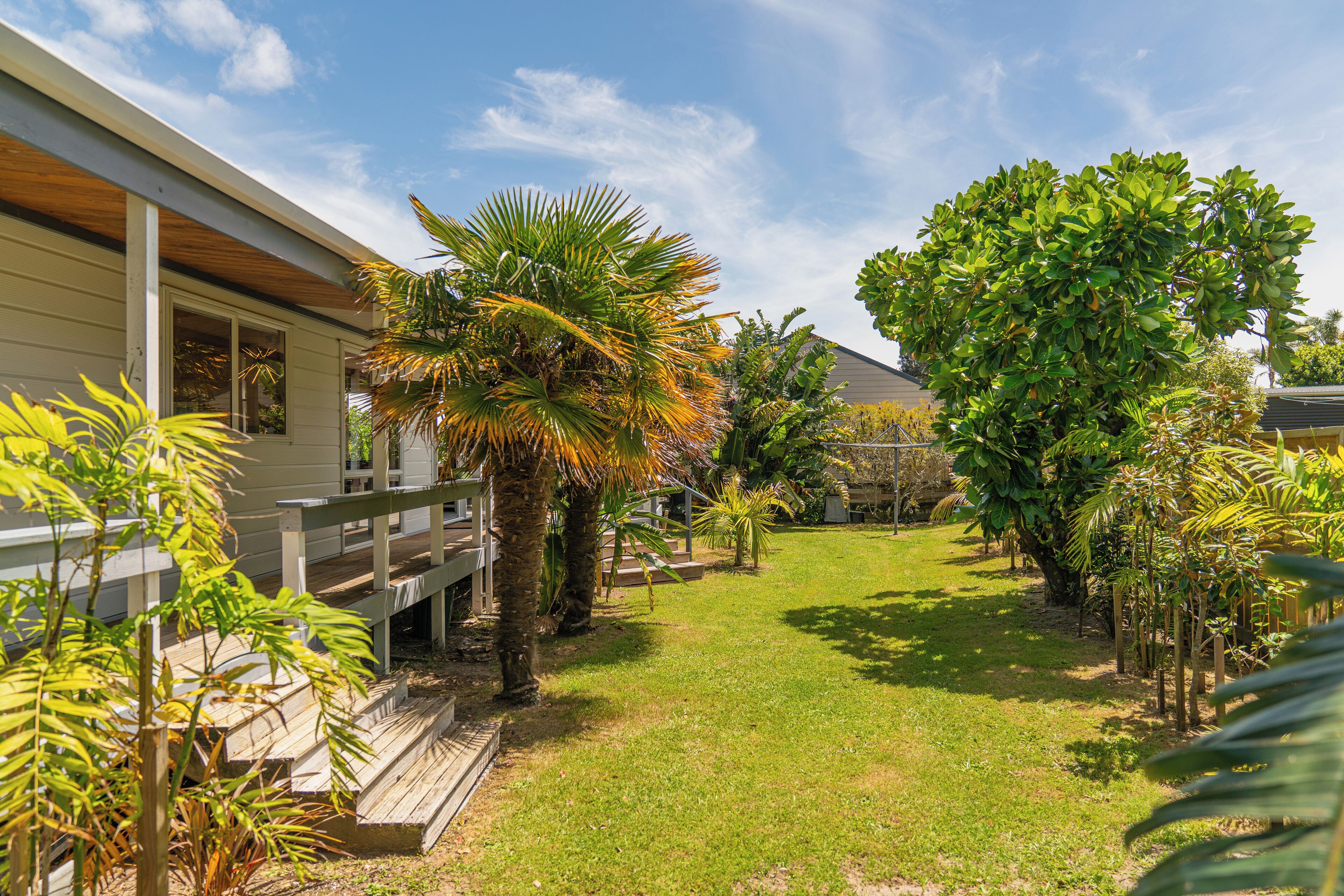 A house with a wooden deck, a grassy yard, and various tropical plants.
