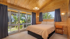 A bedroom with a large bed, wooden walls, and a view of the outdoors. - 5 minutes walk to beautiful beach & shops, fully fenced section pet friendly. (Matarangi)