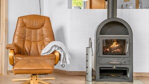 Een moderne woonkamer met een leren fauteuil, een houtkachel en uitzicht op het omliggende landschap door een raam.