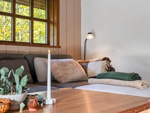 Ein gemütliches Zimmer mit Holztisch, brennender Kerze, einer Kakteenpflanze und Blick auf Bäume durch ein Fenster.
