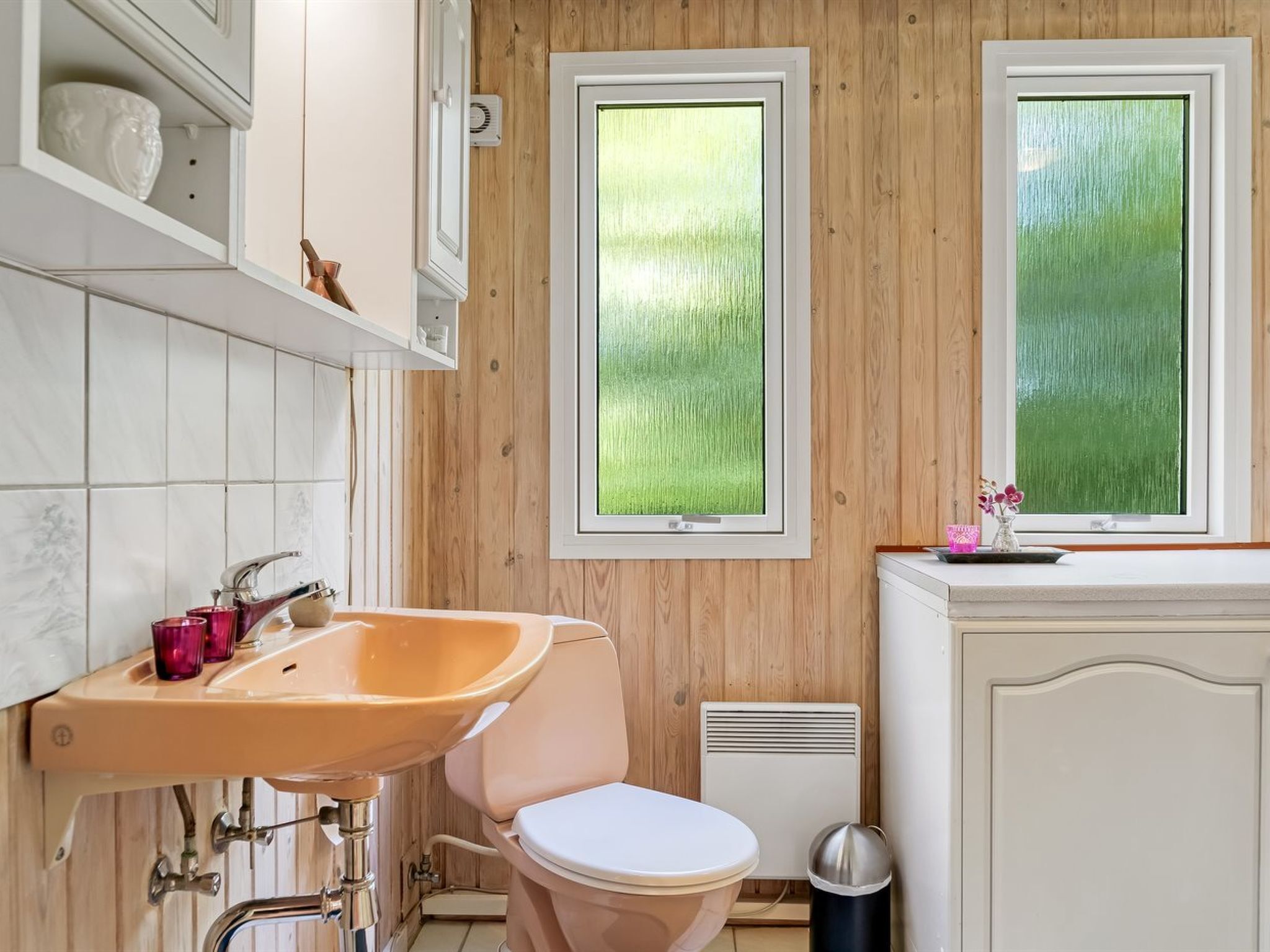 A bathroom with a wooden wall, a white toilet, a beige sink, and a window with a green view.
