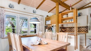 Uma mesa de jantar de madeira com cadeiras, uma luminária pendente e vista para o exterior através de grandes janelas.