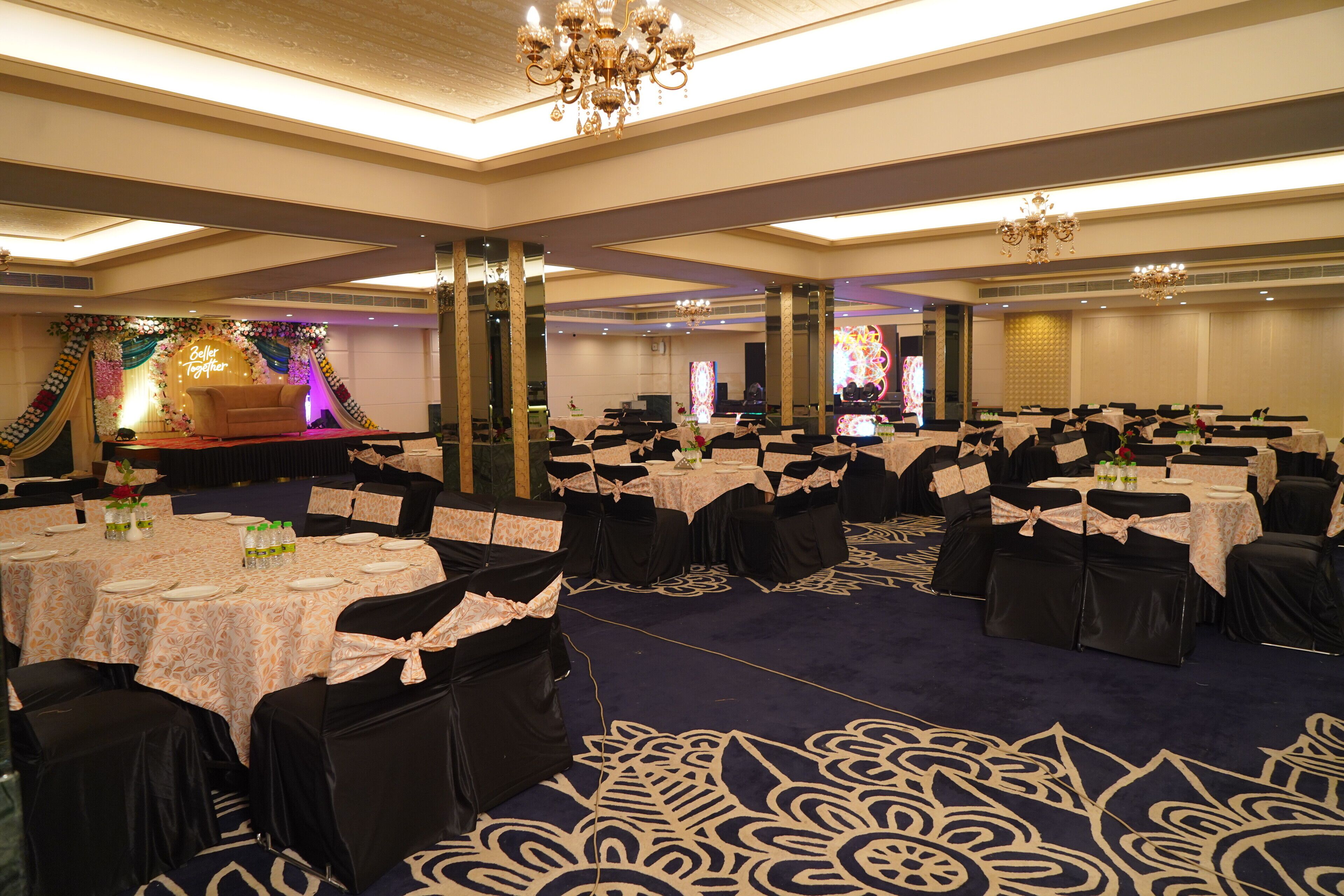 A banquet hall with round tables, black chairs, and a stage with floral decorations.