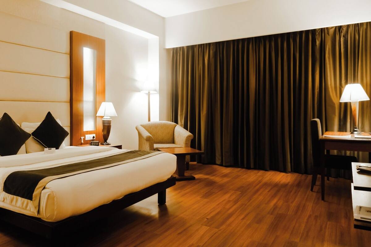 A hotel room with a large bed, a desk, a chair, a lamp, and a large window with curtains.