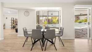 Un comedor moderno con una mesa de vidrio y sillas grises, una estantería y vistas a una piscina a través de amplias ventanas.