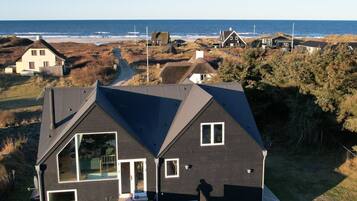 Une maison moderne dotée d’une grande baie vitrée, d’un toit foncé et offrant une vue sur la plage et l’océan.