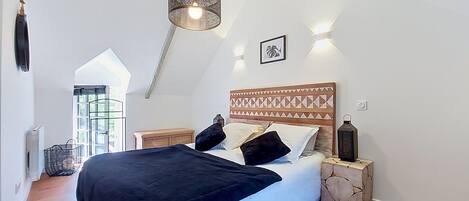Une chambre avec un plafond haut, un lit doté d’une tête de lit à motifs, des lampes de chevet et une fenêtre offrant une vue agréable.
