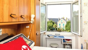 A cozy room with a wooden wardrobe, a bed with a red pillow, a small table with books, and a window offering a view of a house and trees. - Theophile by Interhome (Saint Malo)