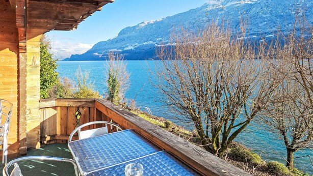 Um deck de madeira com mesa e cadeiras, com vista para um lago e montanhas ao fundo.