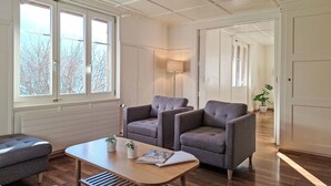 A spacious room with a wooden floor, two grey armchairs, a wooden coffee table, and a large window with a view of bare trees.