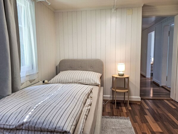 Un dormitorio con cama, mesita de noche con lámpara, suelo de madera y vista a un pasillo.