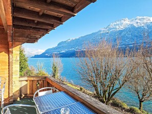 Terraza de madera con una mesa y sillas con vistas a un lago y montañas al fondo.