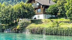 A wooden house with a balcony, surrounded by lush greenery and a body of water.
